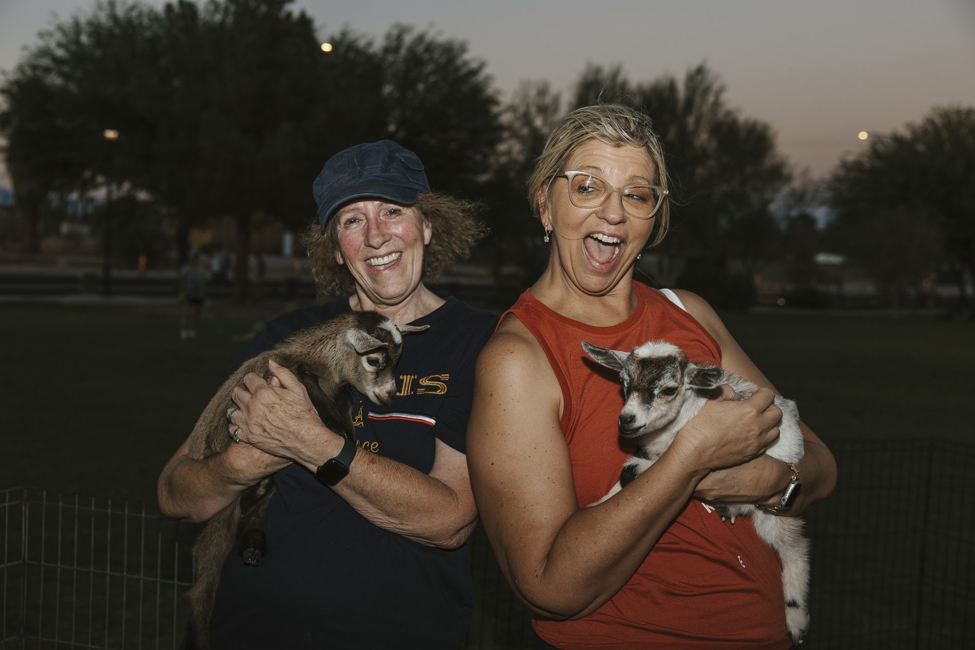 Two women each hold a baby goat and laugh at dusk
